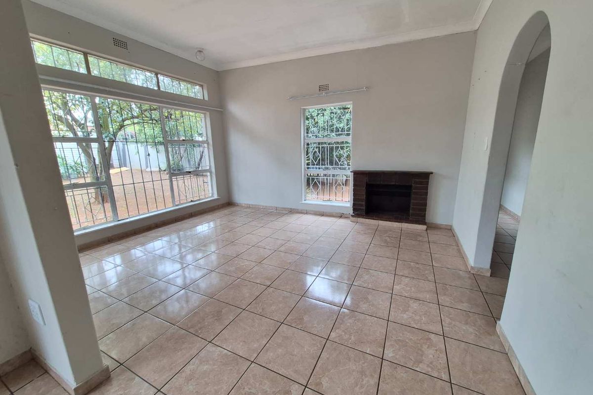 A spacious, empty living room featuring tiled flooring, a brick fireplace, and large windows with security bars, offering a blank canvas for interior design.