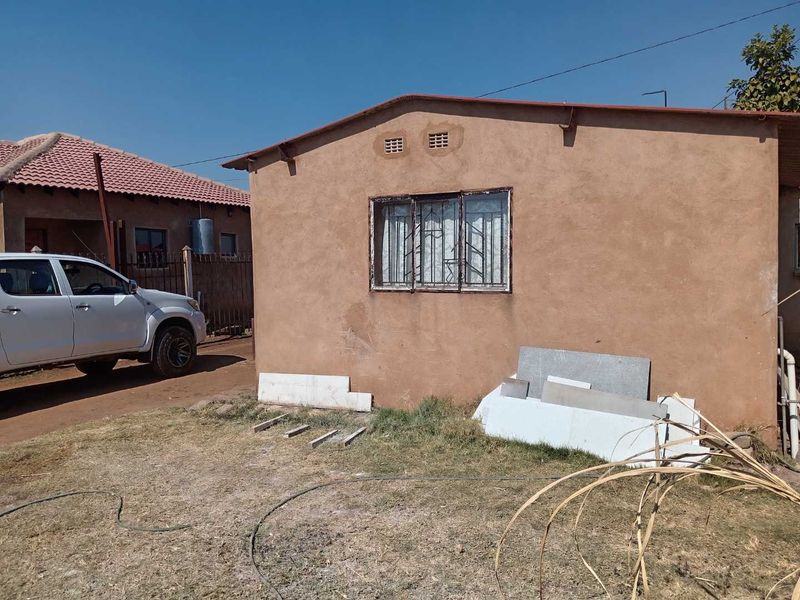 Rear exterior view of the property showing a single-story structure with weathered plaster walls and metal-framed windows with security bars.