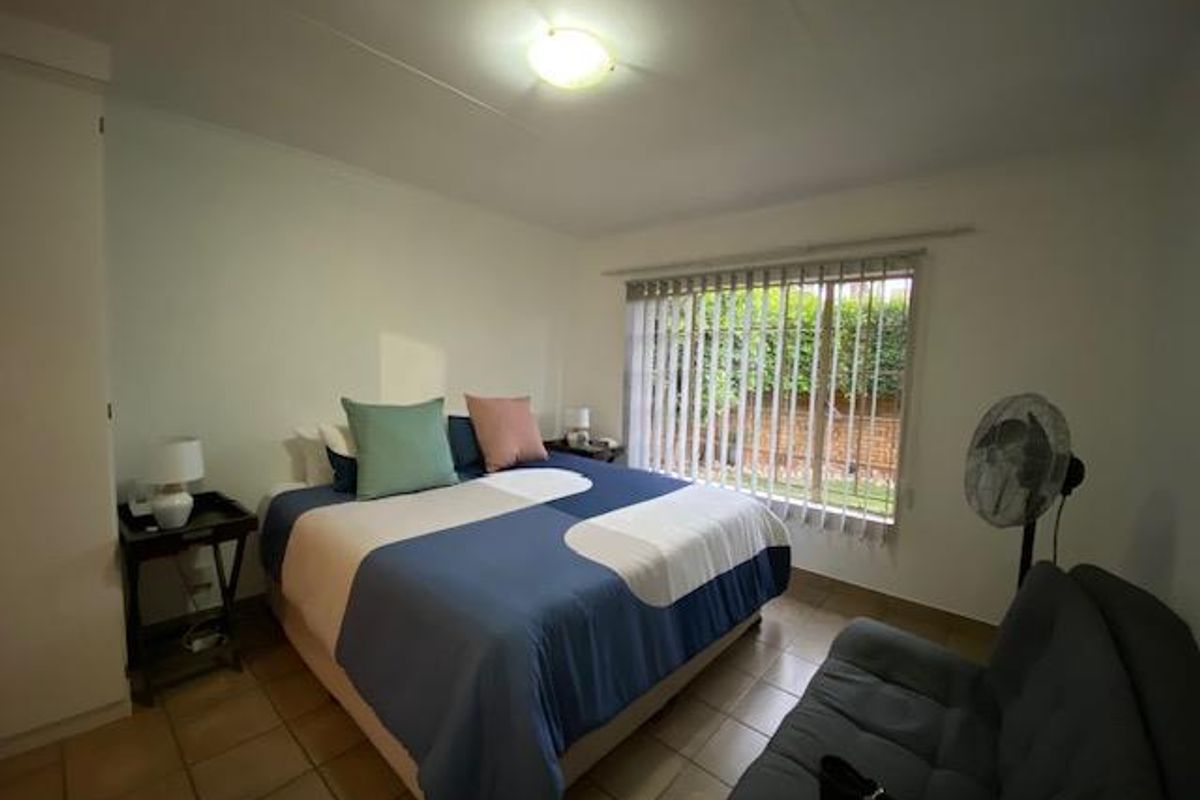 A bedroom featuring tiled flooring, built-in cupboards, and a large window with vertical blinds overlooking the garden.