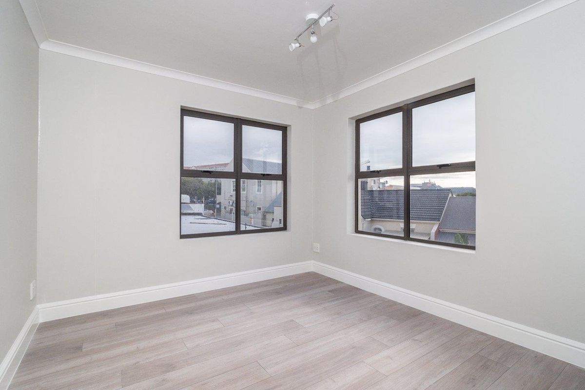 A bright, empty bedroom featuring modern light-toned laminate flooring, neutral walls, and dark-framed windows offering an urban view.