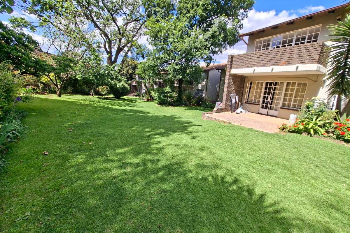 Spacious rear garden featuring a well-maintained lawn, mature trees, and a paved patio area attached to the brick-faced apartment building.