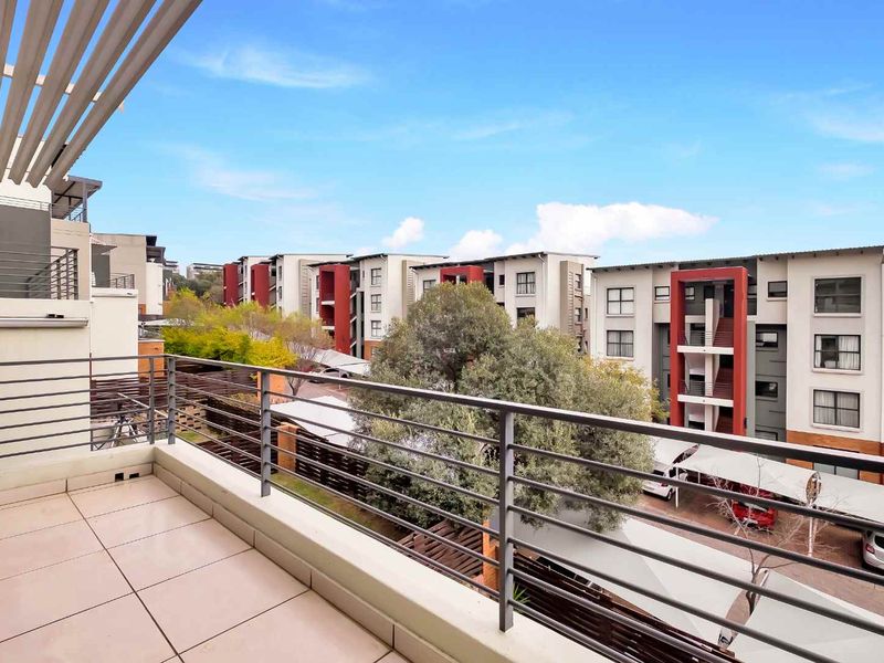 A spacious apartment balcony featuring tiled flooring, a metal balustrade, and a partial pergola, offering views of the surrounding residential complex.