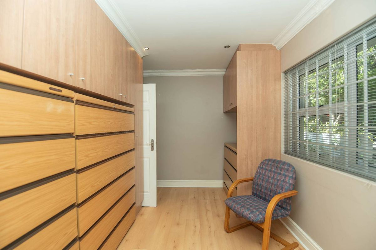 A functional bedroom or dressing area featuring extensive built-in wooden cabinetry, laminate flooring, and natural light through a window with fitted blinds.
