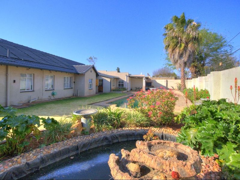 Rear garden view featuring a swimming pool, paved patio area with built-in braai, and a landscaped pond in the foreground.