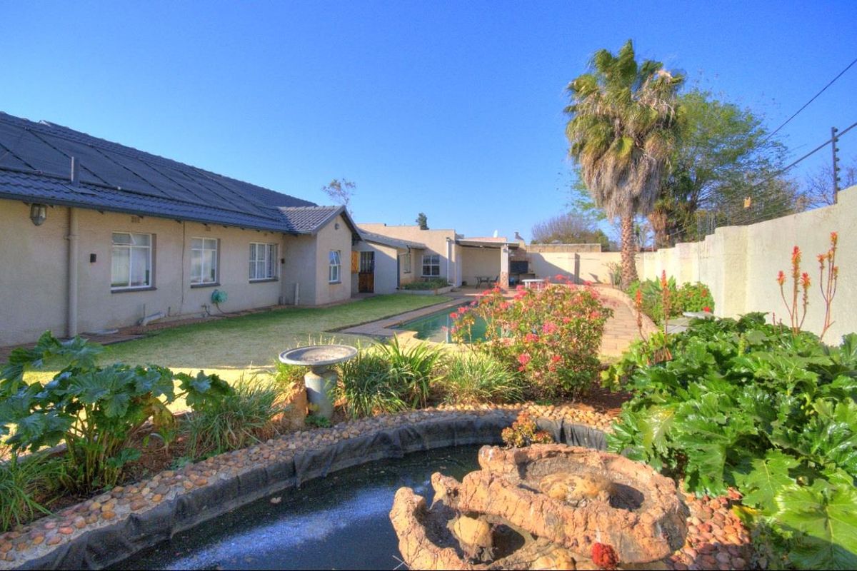 Rear garden view featuring a swimming pool, paved patio area with built-in braai, and a landscaped pond in the foreground.
