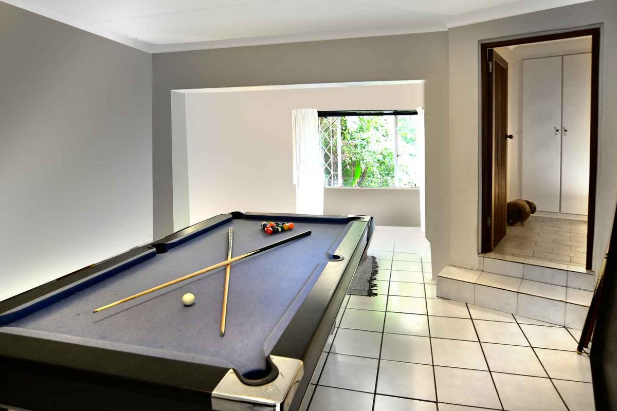 A spacious living area featuring tiled flooring, a raised step leading to a hallway with built-in cupboards, and large windows overlooking the garden.