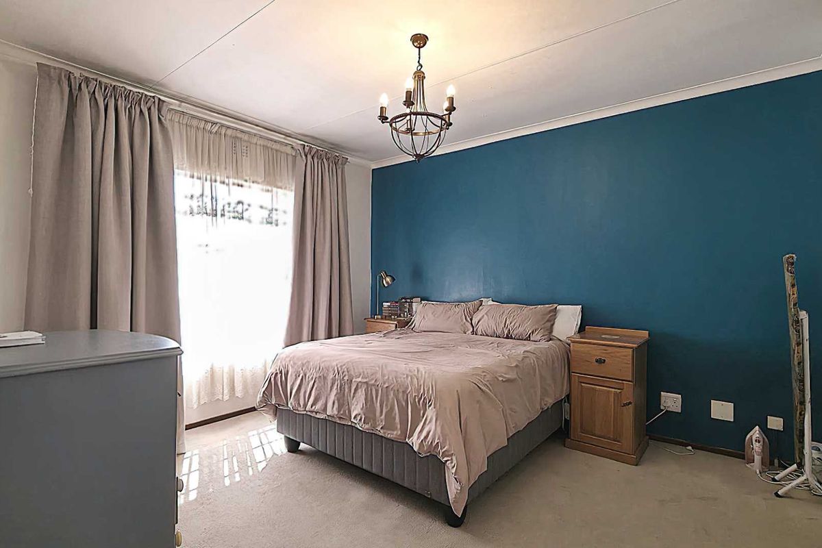 A well-lit bedroom featuring a prominent blue accent wall, neutral carpeting, and a central ceiling light fixture.