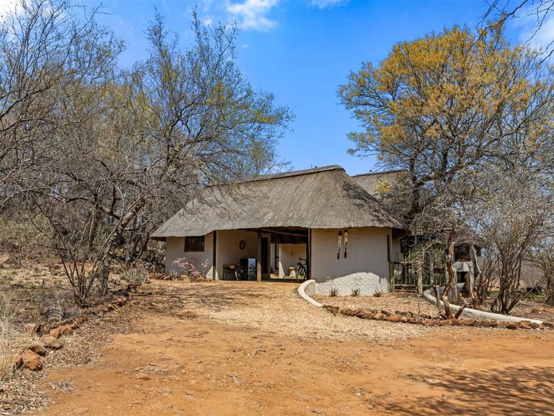A charming bushveld-style home featuring a traditional thatched roof and a natural gravel driveway, set within a scenic, wooded landscape.