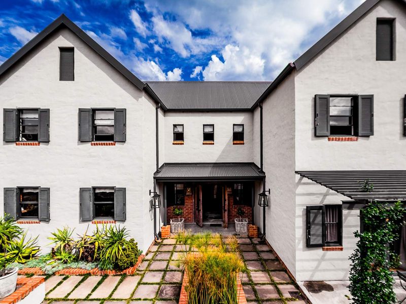 A well-maintained two-story residence featuring a classic architectural design with white rendered walls, dark metal roofing, and a paved front courtyard entrance.