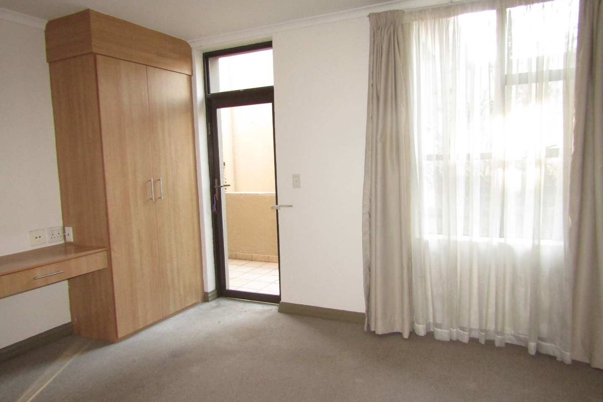 A bright, empty bedroom featuring built-in cabinetry, a desk, carpeted flooring, and direct access to a private balcony through a glass door.