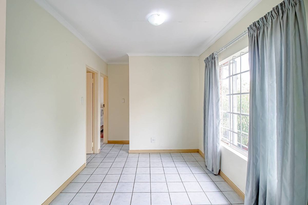 A bright, empty living area featuring neutral tiled flooring, large window with security bars, and access to adjacent rooms.