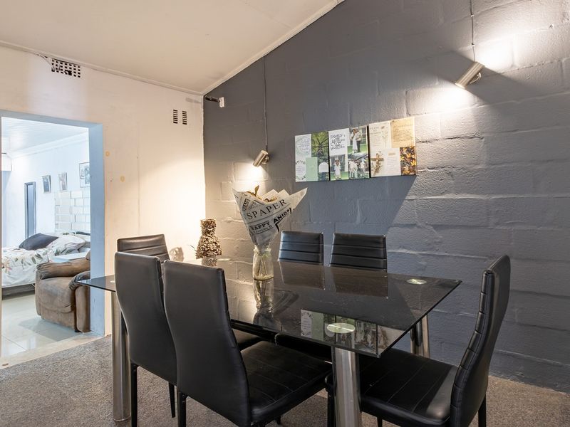 Dining area featuring a dark painted exposed brick feature wall, carpeted flooring, and modern spotlighting, with an adjacent view into a bedroom.