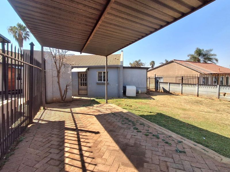 Rear exterior view featuring a paved patio area, a corrugated metal carport, and secure perimeter fencing surrounding the garden space.