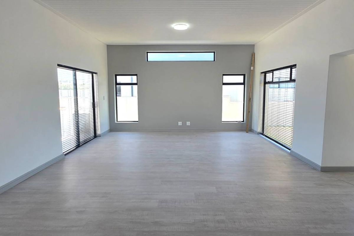 A spacious, light-filled living area featuring modern grey laminate flooring, neutral walls, and large windows with fitted blinds.