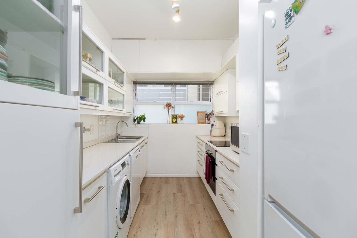 A bright, galley-style kitchen featuring white cabinetry, integrated appliances, a stainless steel sink, and light wood-look flooring.