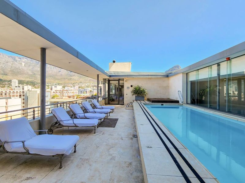 A rooftop pool area featuring a modern swimming pool, stone tiling, and a covered patio section with panoramic mountain and city views.