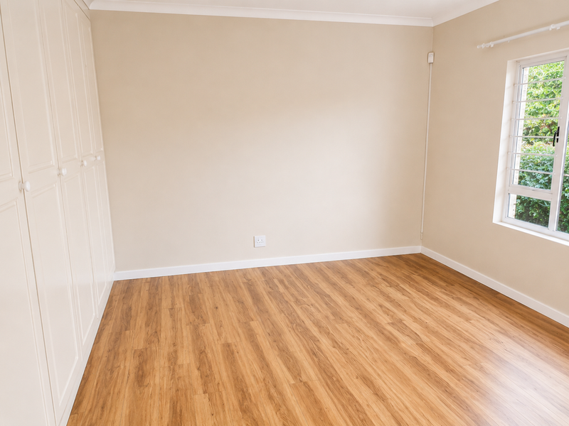 A bright, empty bedroom featuring light-toned laminate flooring, neutral walls, and ample built-in cupboard storage with a window overlooking the garden.