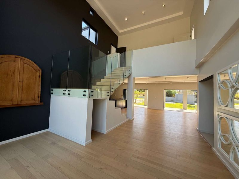 Spacious open-plan living area featuring modern timber flooring, a striking glass-railed staircase, and double-volume ceilings with recessed lighting, leading out to the garden.