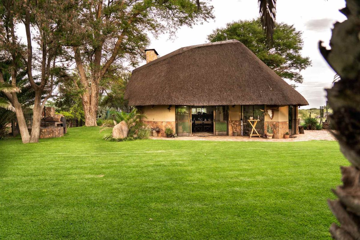 A charming thatched-roof farm cottage set within a lush, well-maintained garden, featuring stone cladding and a built-in outdoor braai area.