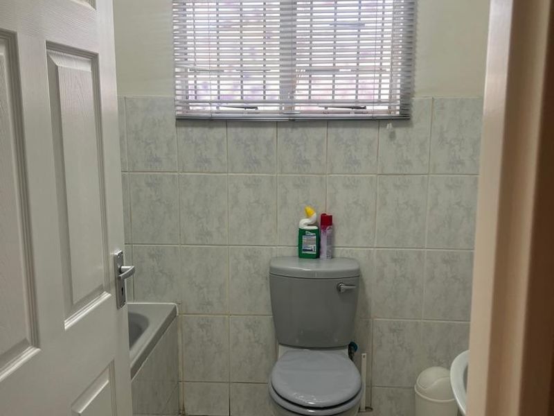 A functional bathroom featuring tiled walls and flooring, a standard toilet, and a bathtub with a window fitted with venetian blinds.