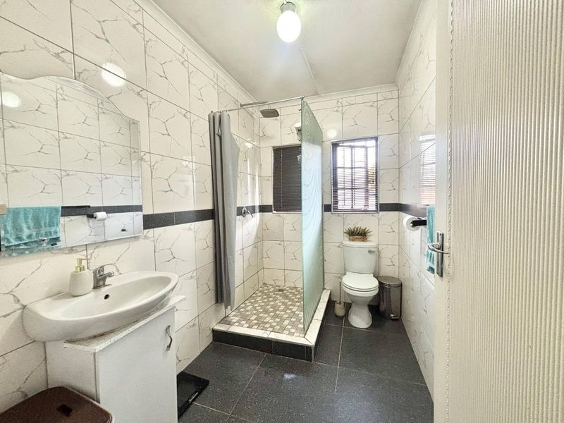A functional bathroom featuring a walk-in shower with a glass screen, vanity basin, and toilet, finished with marble-effect wall tiles and dark floor tiles.