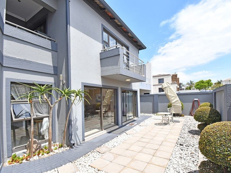Rear exterior view of the property featuring a paved patio area, sliding glass doors, and an upper-level balcony with modern grey facade finishes.