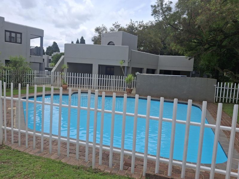 A well-maintained communal swimming pool area featuring secure white fencing, brick paving, and surrounding landscaped grounds.