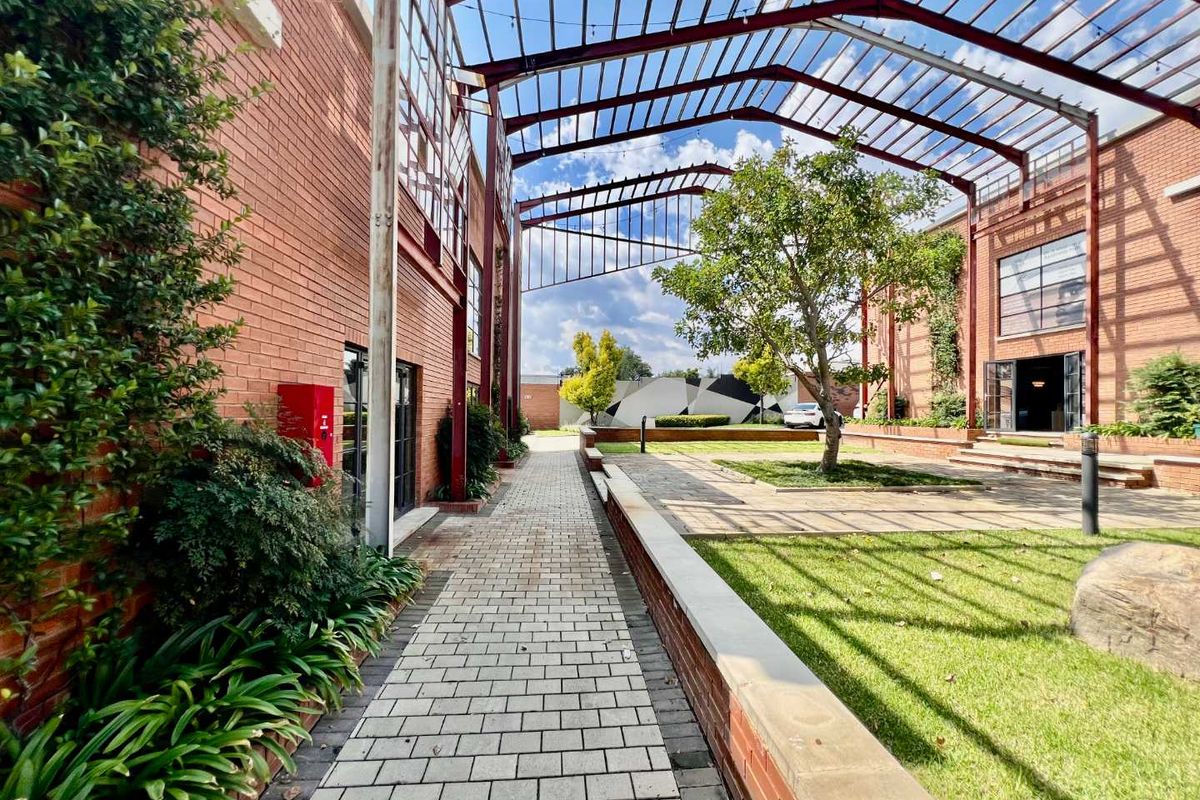 A well-maintained outdoor courtyard featuring brick pathways, landscaped garden beds, and a prominent steel pergola structure connecting the business premises.