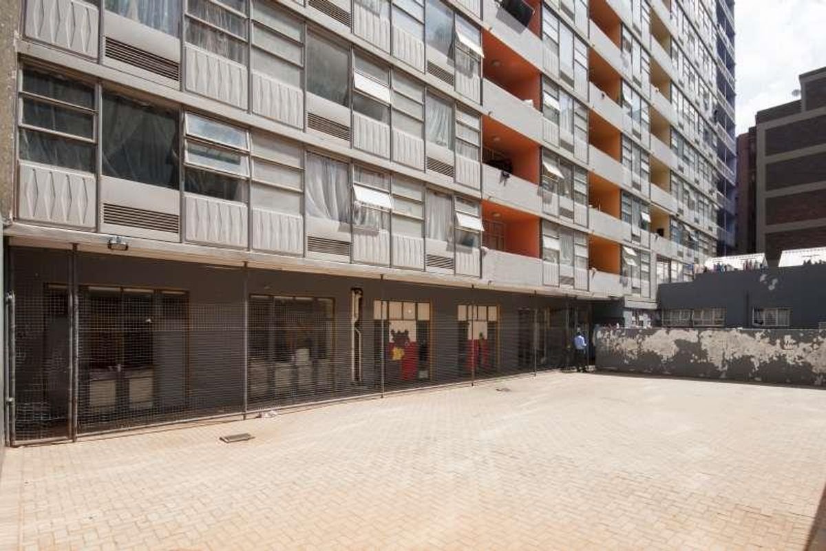 Exterior view of a multi-story apartment building featuring a paved courtyard area and security fencing at the ground level.