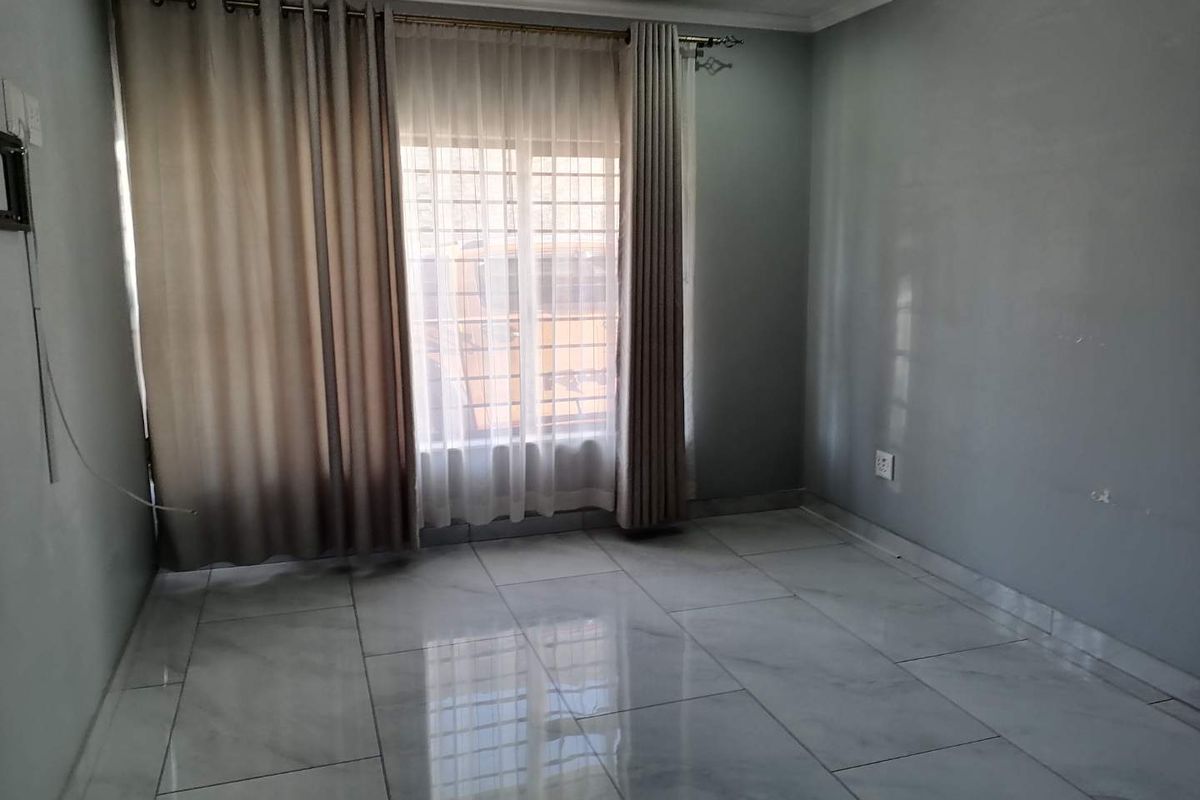 A bright, empty bedroom featuring polished tiled flooring, neutral grey walls, and large windows fitted with security bars and curtain tracks.