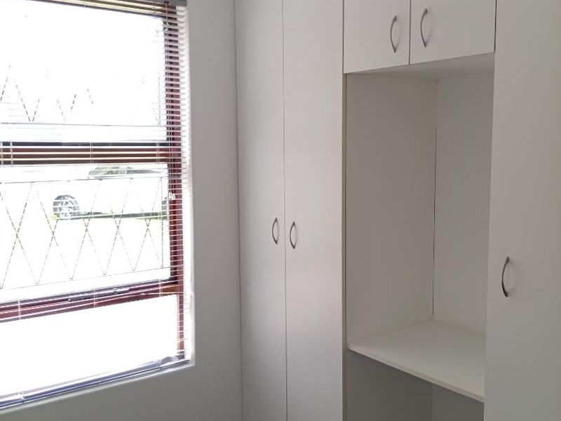 A clean, empty bedroom featuring white built-in cupboards with an integrated desk area and a window fitted with security bars and blinds.