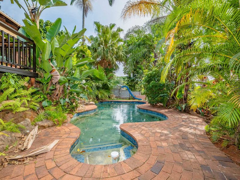 A backyard swimming pool area featuring brick paving, lush tropical landscaping, and a built-in pool slide.