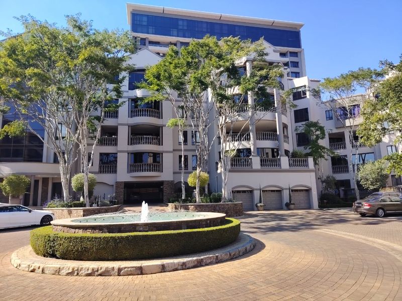 Front elevation of a multi-story apartment complex featuring a circular driveway, central fountain, and structured balconies with stone balustrades.