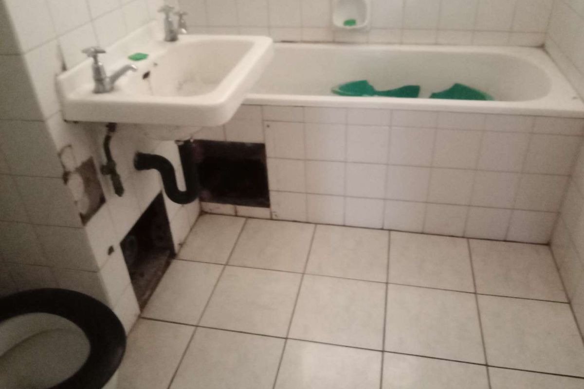 A dated bathroom featuring a standard bathtub, wall-mounted basin, and toilet, currently requiring significant repairs to wall tiling and exposed plumbing infrastructure.