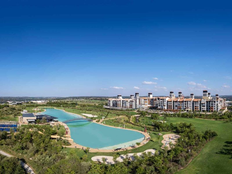Aerial view of a residential estate featuring a large man-made lagoon, pedestrian bridge, sports facilities, and extensive landscaped green spaces.
