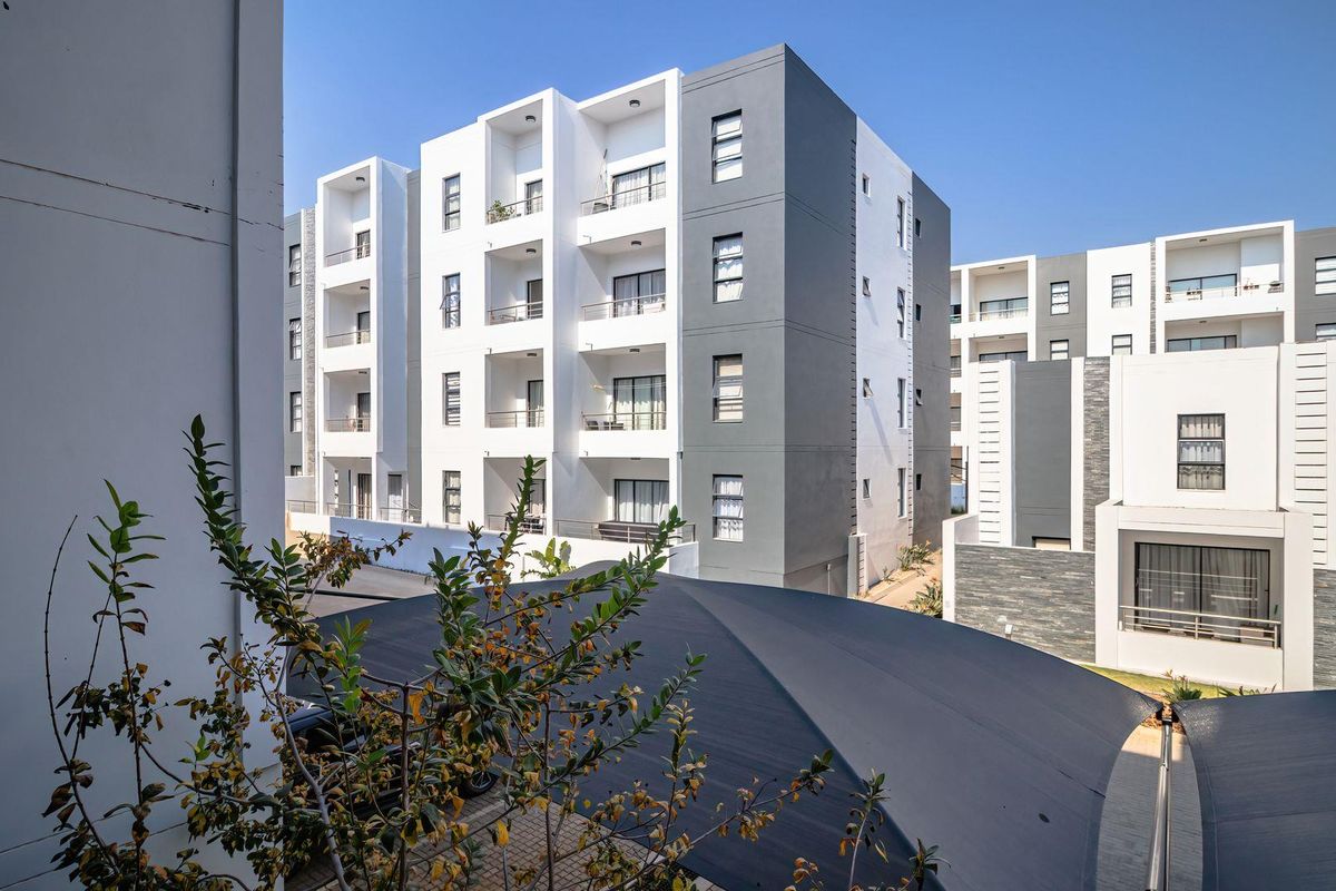 A view of a modern multi-story apartment complex featuring clean architectural lines, private balconies, and a paved parking area with shade sails.