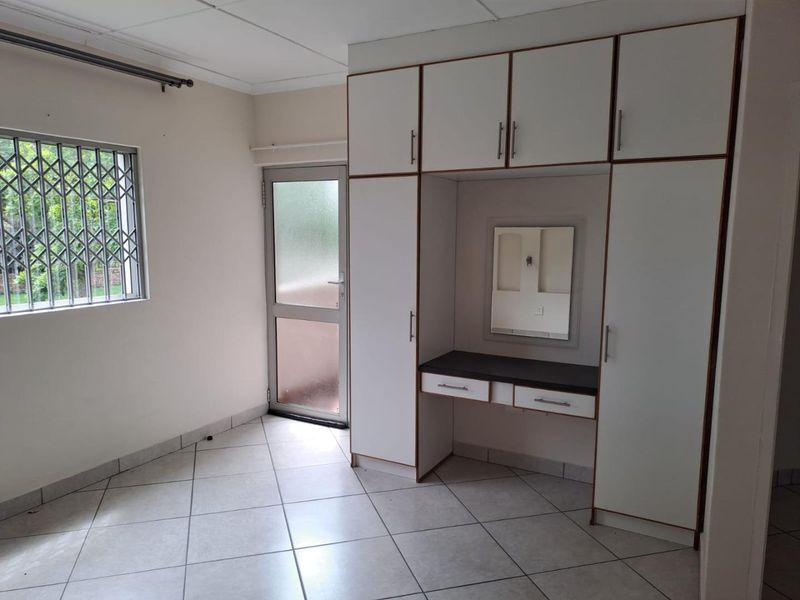A clean, empty bedroom featuring built-in cupboards with an integrated vanity station, tiled flooring, and direct access to an exterior door.
