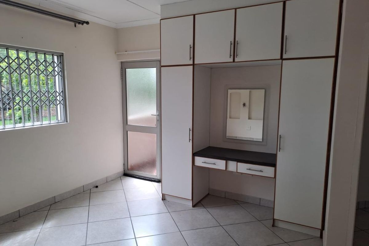 A clean, empty bedroom featuring built-in cupboards with an integrated vanity station, tiled flooring, and direct access to an exterior door.