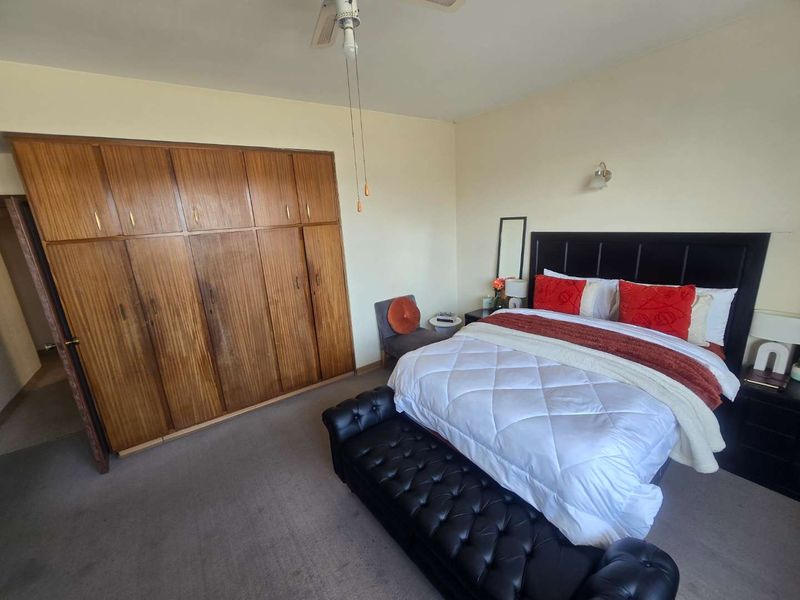 A spacious bedroom featuring a large built-in wooden wardrobe, neutral wall finishes, and a ceiling fan, offering a functional layout with carpeted flooring.