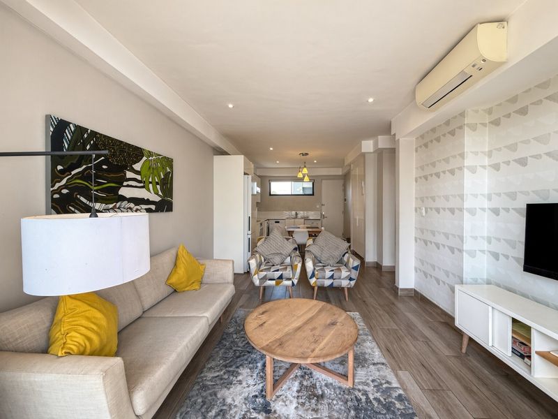 A bright open-plan living and kitchen area featuring modern laminate flooring, recessed lighting, a split-system air conditioner, and a stylish feature wall.