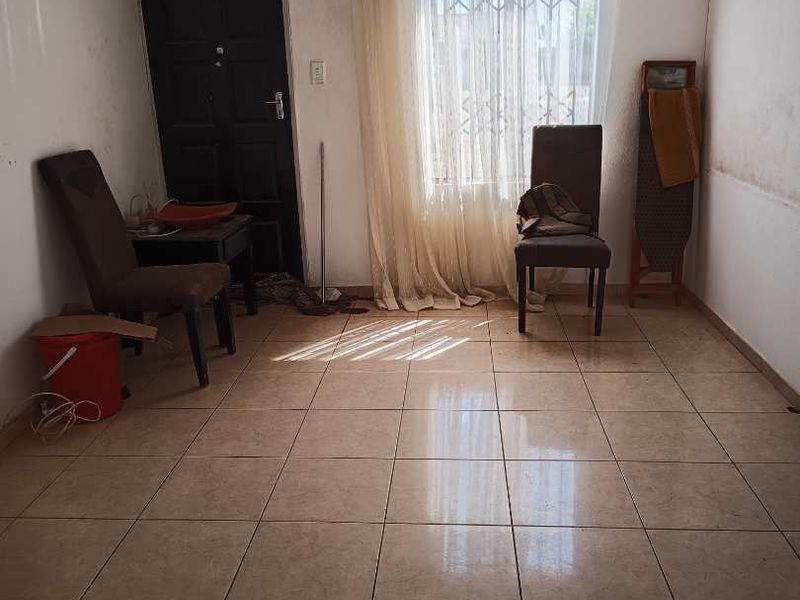 A sparsely furnished living area featuring tiled flooring, a security gate, and natural light through a window with curtain rails.