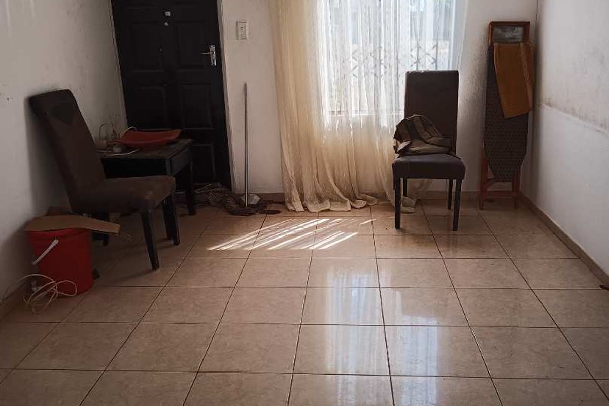 A sparsely furnished living area featuring tiled flooring, a security gate, and natural light through a window with curtain rails.