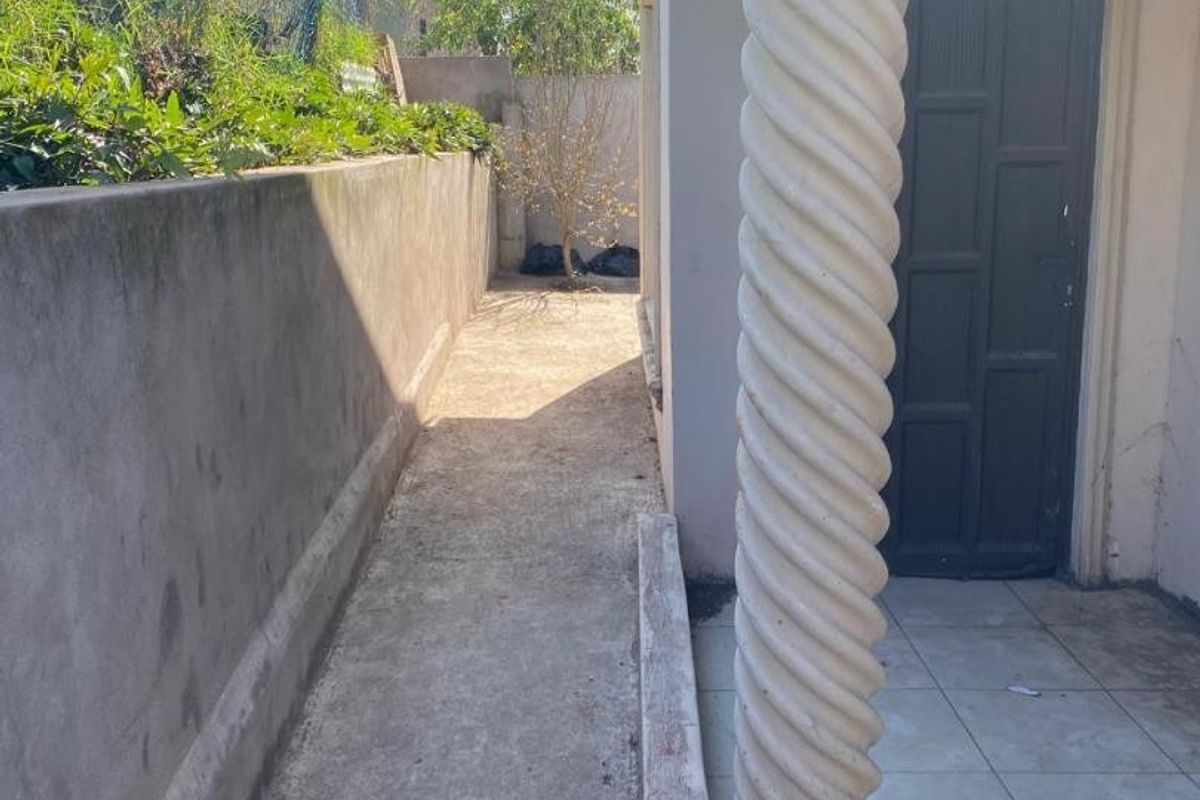 A narrow exterior side passage featuring a concrete walkway, a decorative column, and a tiled porch area, requiring maintenance and surface repairs.