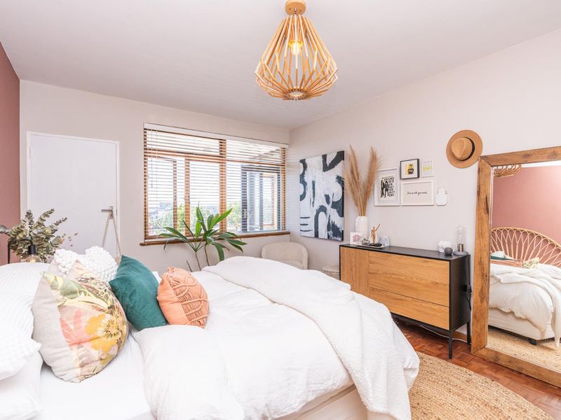 A well-lit bedroom featuring warm parquet flooring, a large window with venetian blinds, and a modern pendant light fixture.
