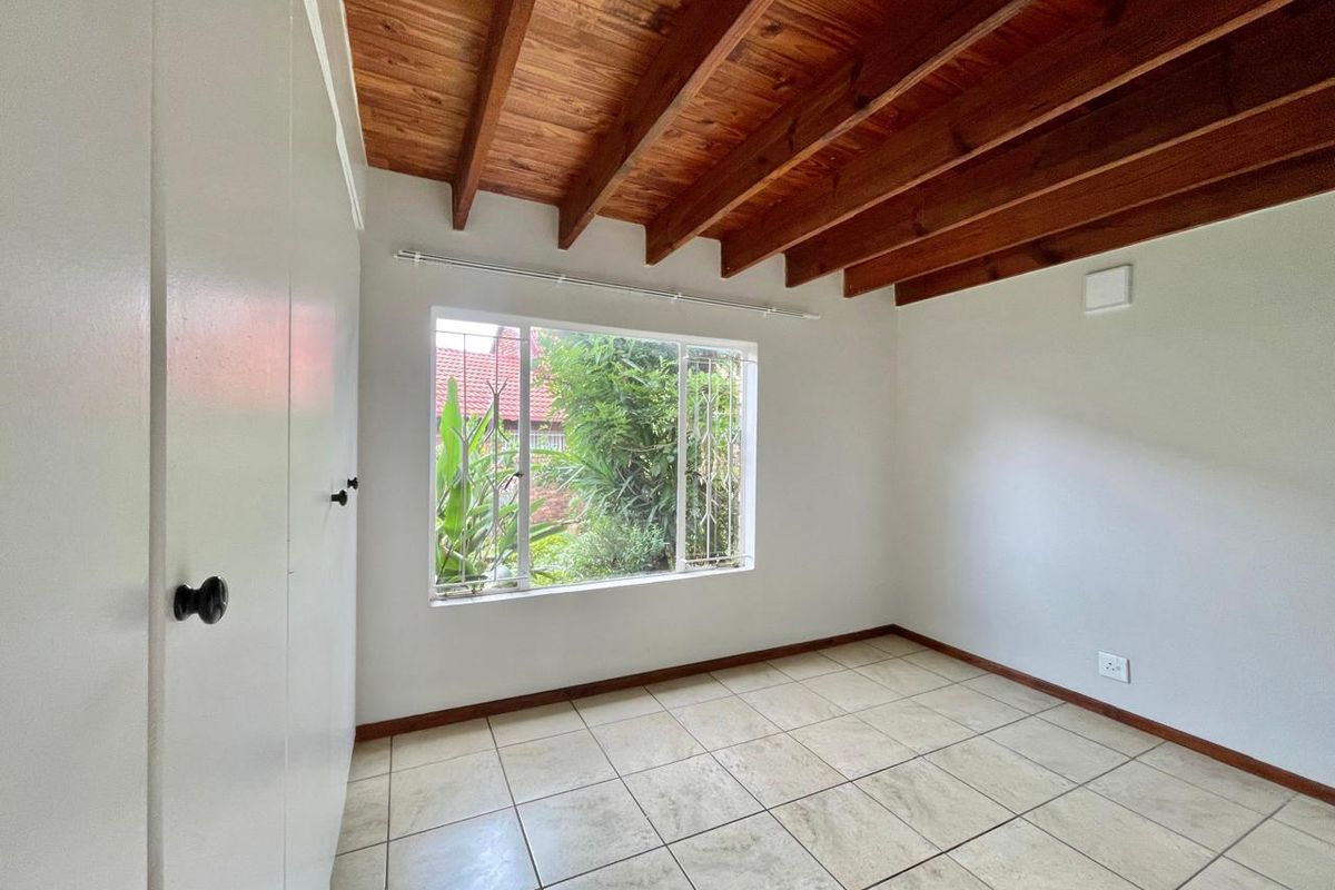 A bright, empty bedroom featuring exposed timber ceiling beams, tiled flooring, and built-in cupboards with a view of the garden.