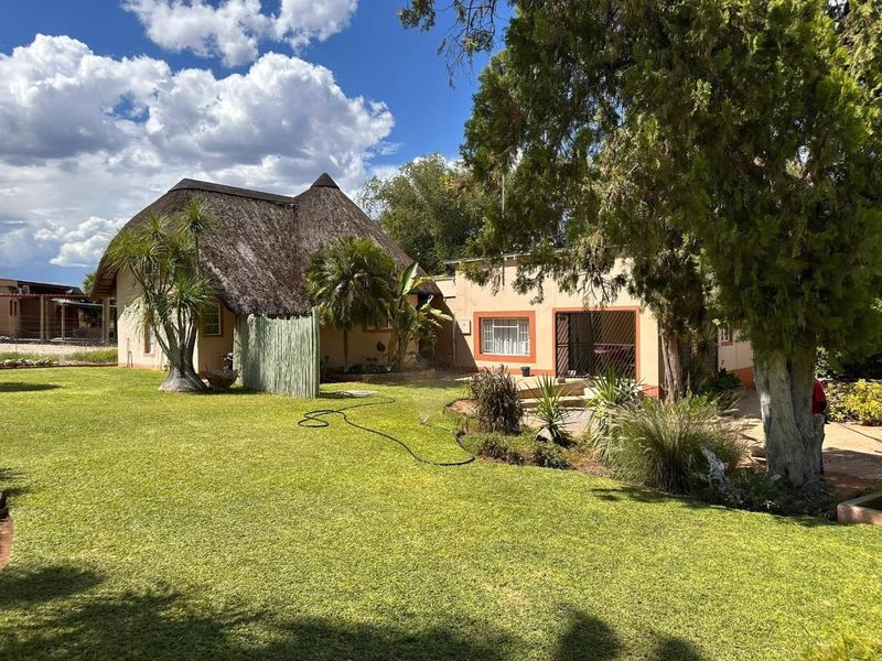 A view of the rear garden featuring a traditional thatch roof structure, paved patio area, and established trees on a well-maintained lawn.