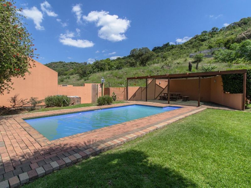 A well-maintained outdoor pool area featuring a rectangular swimming pool with brick coping, a timber pergola, and a surrounding lawn set against a hillside.