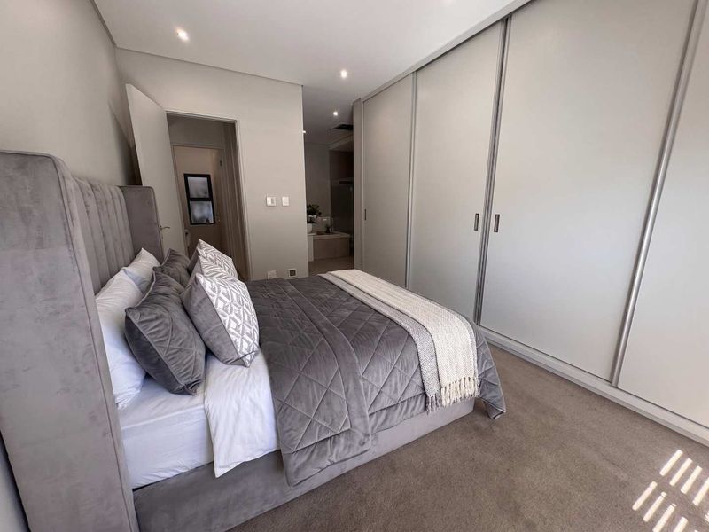 A modern bedroom featuring neutral tones, plush carpeting, and extensive built-in floor-to-ceiling cupboards, with an adjoining en-suite bathroom visible.