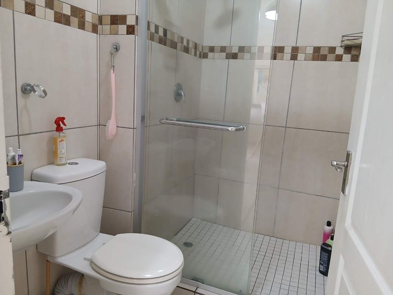A functional bathroom featuring a glass-enclosed shower, pedestal basin, and neutral wall tiling with a decorative mosaic border.