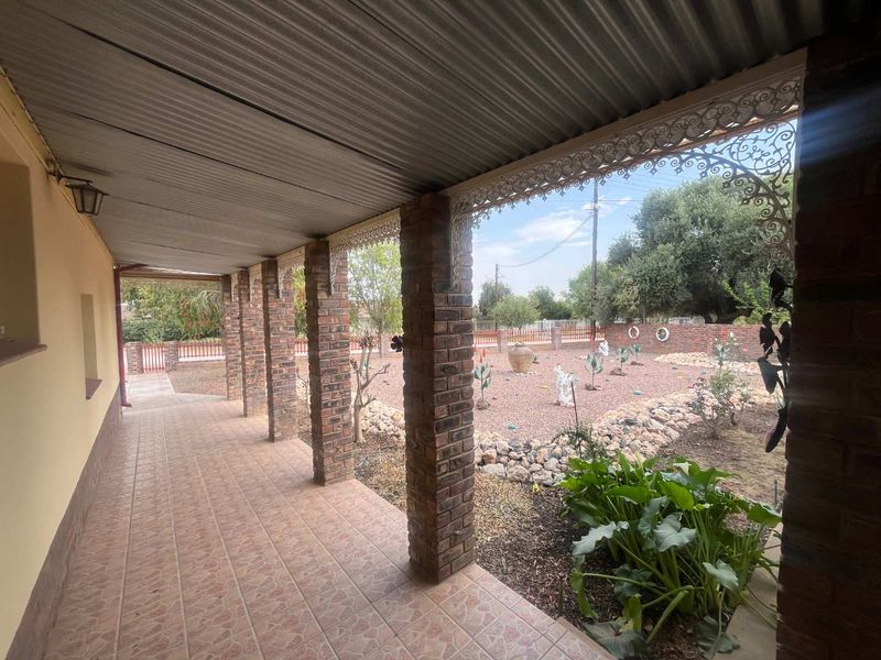 A covered patio featuring brick pillars, decorative wrought iron detailing, and tiled flooring, overlooking a landscaped garden area with a brick boundary wall.